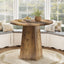 Wooden dining table with a round top and a single pedestal base in a room with large windows.